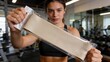 © ibhonk - Athletic woman with toned physique holds beige resistance band packaged in clear plastic inside modern gym