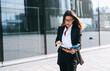 © Iona - Smiling business professional outside a sleek modern building, holding a phone and folder, radiating confidence and readiness.