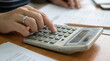 © paulmalaianu - Close-up of a woman's hand using a calculator to manage finances, calculate taxes, or budget at a wooden desk with documents, 3D illustration.