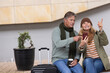 © wavebreak3 - Senior couple sitting on plaza bench wearing scarves holding coffee, checking phone near suitcase
