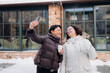 © m-art - Two friends stand together in a snowy setting, laughing and taking selfies. They are dressed warmly, holding cups, and the brick wall gives a charming backdrop to their joyful moment.