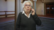 © Krakenimages.com - Senior woman holds smartphone to ear with visible hand and glasses in a building foyer; concern aging.
