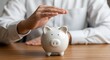 © PIXEL BOY - Person protecting a white piggy bank with their hand on a wooden table