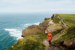 © Agnieszka Gaul - A person in a bright orange jacket walks along a narrow coastal path winding high above the churning blue waters of the Atlantic at the Cliffs of Moher.