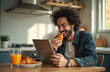 © Maryna - Man eats croissant at kitchen table while watching tablet. Orange juice and pastry sit nearby. He laughs enjoying morning content, browsing or searching online. He wears casual attire indoors.