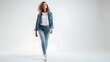 © faisal - A confident young female with curly blonde hair walks forward wearing a casual blue denim jacket over a white shirt and fitted blue jeans with white canvas sneakers. She poses against