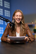 © fizkes - Portrait of smiling female host sitting at studio desk, holding script or notes papers while speaking into mounted broadcast microphone. Content creation for podcast, online show, or radio program