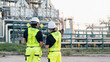 © AU USAnakul - Two men in bright yellow vests and white helmets inspect large industrial plant with pipes and structures, conducting safety review and equipment assessment under clear sky, examining refinery.