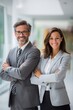 © The Little Hut - Confident business colleagues, a man and woman in suits, standing with arms crossed in a modern office hallway, exuding professionalism.