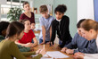 © JackF - Group of teenagers and students discuss the task at the table, they exchange opinions and theories against the background of teacher in classroom. Schoolchildren look at the paper and solve the task