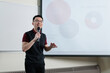 © fotolite - A smiling Asian man wearing glasses holds a microphone while gesturing during a presentation in front of a chart, ideal for business pitch, school presentation, or public speaking concepts.