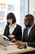 © sofiko14 - A woman in glasses and a man in a suit collaborate on a project at a desk with a laptop and charts on the wall