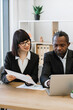© sofiko14 - A woman and an African American man in business attire collaborate over documents and a laptop at a desk in a modern office setting