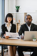 © sofiko14 - A woman and an African American man in business attire sit at a desk with a laptop and documents, looking towards the camera