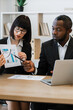 © sofiko14 - A woman in glasses points to a financial chart while discussing with an African American man at a desk with a laptop