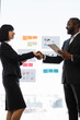 © sofiko14 - Two professionals, an African American man and a woman, shake hands in front of a glass wall with charts and sticky notes, symbolizing a successful deal
