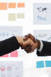 © sofiko14 - Two professionals, one African American man and one woman, shake hands in a gesture of agreement and partnership in a modern office setting with charts and sticky notes visible