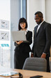 © sofiko14 - A woman and an African American man in suits collaborate over a laptop in a modern office setting with charts on the wall