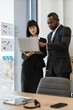 © sofiko14 - A focused African American man and a woman in business attire collaborate over a laptop, reviewing charts and data on a glass wall