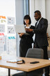 © sofiko14 - A focused African American man and a woman in business attire collaborate over a laptop, reviewing charts and graphs displayed on a window