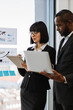 © sofiko14 - A woman and an African American man in business attire review data on a laptop and tablet near a window with charts on the wall