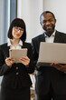 © sofiko14 - A smiling African American man holds a laptop while a woman with glasses holds a tablet, both dressed in business attire
