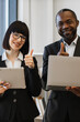 © sofiko14 - A smiling woman and an African American man in business attire give a thumbs up while holding a tablet and laptop, signifying approval and achievement