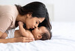 © theshooter - Madre joven besando con ternura a su bebé tumbado sobre una cama con luz natural, concepto de amor maternal, maternidad, cuidado y salud infantil para publi de lifestyile y seguro de vida