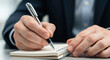 © julijadmi - Business professional writing notes in a spiral notebook with a silver pen on a clean desk surface
