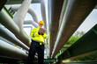 © FotoArtist - Industrial worker in yellow jacket and hard hat using tablet near large pipes