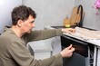 © pundapanda - A man stands in his kitchen taking clean dishes out of the dishwasher. He focuses on the process of unloading the clean dishes and organizing them for use