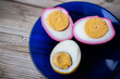 © Jolanta - Close-up of halved pickled eggs with vibrant pink and yellow whites on a dark blue plate, showing firm yolks and shallow depth of field. Soft background blur.