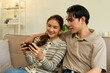 © MIND AND I - Happy Young Asian Couple Watching Smartphone Together on Sofa at Home