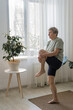 © ximich_natali - Elderly woman balancing on one leg, stretching and doing fitness in her lounge with houseplants, staying active for healthy aging, exercising at home, practicing yoga wellness