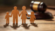 © tl6781 - Wooden cutout figures of a family stand in front of a blurred judge’s gavel. The scene suggests themes of family law, custody, or legal decisions affecting households