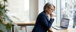 © Yuliia - Mature businesswoman with glasses working on laptop in modern office, looking thoughtful while reviewing data by window with city view.