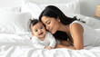 © theshooter - Madre joven con ternura junto a su bebé tumbado sobre una cama con luz natural, concepto de amor maternal, maternidad, cuidado y salud infantil para publi de lifestyile y seguro de vida