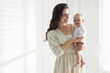 © New Africa - Smiling mother in dress holding her cute baby near white wall at home. Space for text