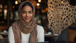 © demzp - Young Muslim woman wearing a hijab smiles warmly while holding a tablet in a cafe