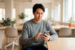 © designX - Focused Asian man using smartphone in modern bright office workspace while sitting at desk during break or remote work