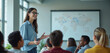 © Viktor - Professor talks to diverse students in university classroom. Young woman with glasses presents lecture with hand gestures. People listen attentively at desks in modern school hall.