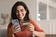© fizkes - Happy Latina woman sits in living room with card and smartphone, making online purchase, paying bill through banking app, managing subscription services, sending money via mobile payment application