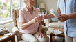 © solom - elderly woman receiving glass of ice water and portable handheld cooling fan from caregiver at home during summer heatwave for hydration and relief from high temperatures