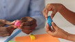 © Prostock-studio - Two hands are shaping pieces of clay at a table. One hand holds a blue piece while the other has a pink piece. They are engaged in a creative activity, surrounded by colored paper and tools.