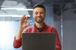 © Prostock-studio - A man is smiling and waving while participating in a video call from a modern office. He is focused on the screen and appears to be enjoying the conversation.