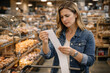 © Khemmanat - Price Check Predicament: A woman examines a long receipt in a brightly lit market, expressing concern, as she contemplates the cost of goods and groceries.