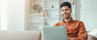 © Prostock-studio - A man sits on a couch working on a laptop. He smiles while looking at the screen. A bright room with shelves and books is in the background. Natural light fills the space.