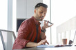 © Prostock-studio - A man is sitting at a desk in an office. He is using a voice recorder while focusing on notes in a notebook. A laptop and a cup are on the table, and sunlight is coming through the window.