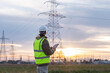 © 24K-Production - A young male engineer checks high-voltage power lines with a tablet outdoors during sunset, focusing on safety and energy management.