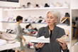 © JackF - Senior female customer is standing next to shelf in stores sales area. She picks up snickers, looks at and studies material and quality of product, decides to buy shoes